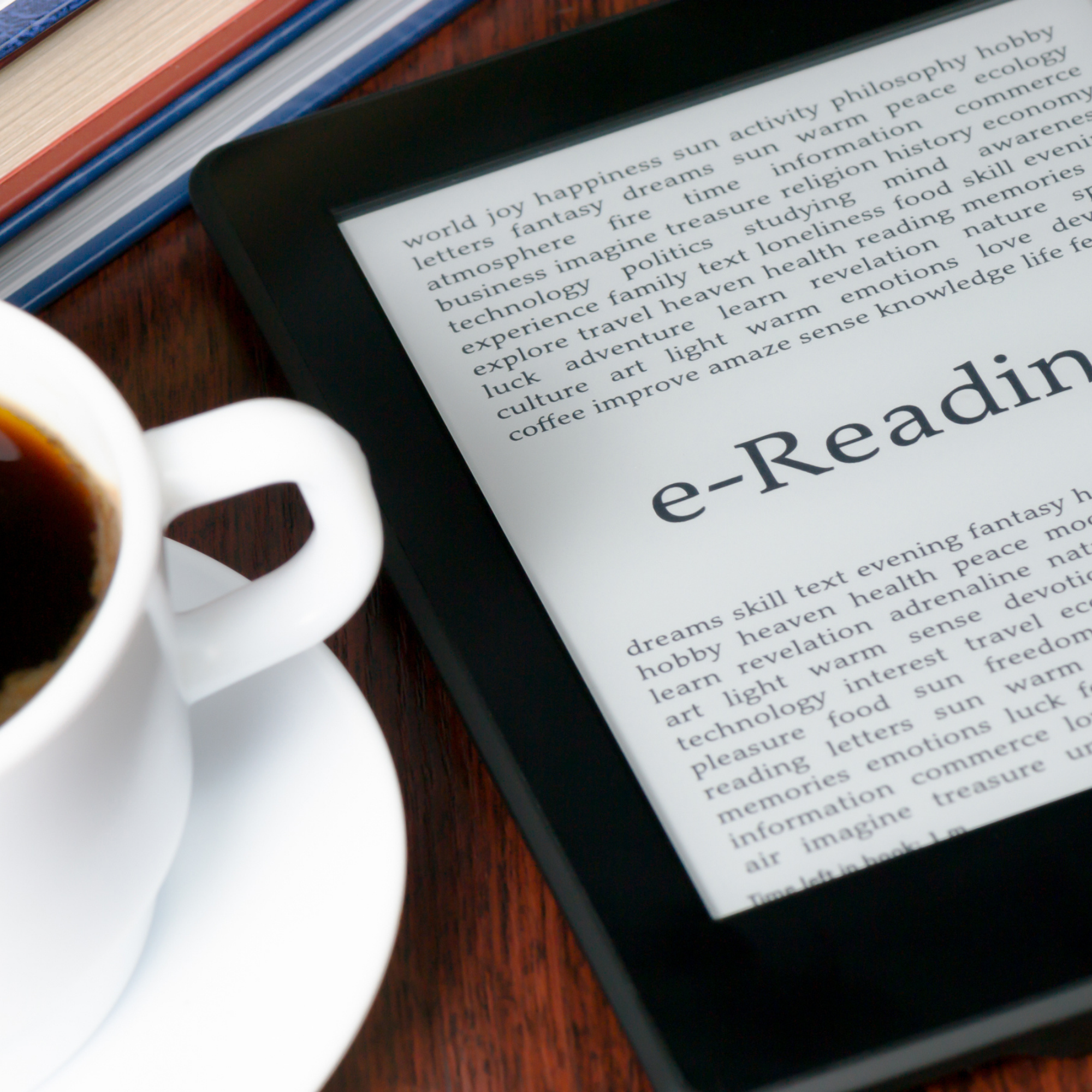 There is a white coffee cup with full of coffee out of frame on the left, and on the right is an e-reader device. 