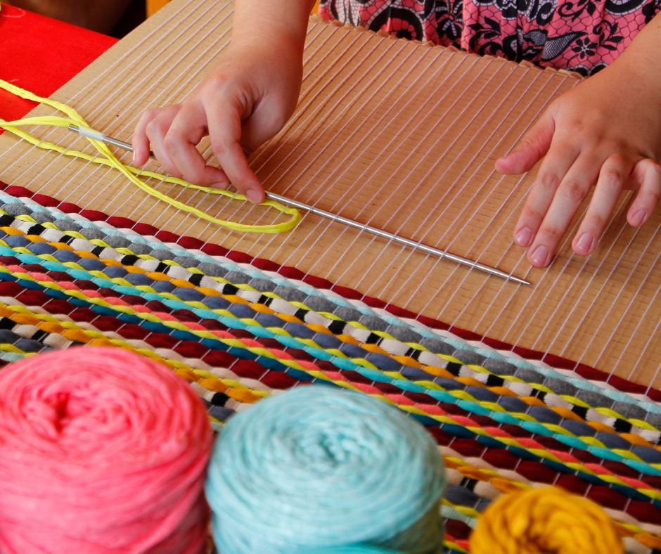 a cardboard loom, with hands weaving a thread through it.  A few skeins of yarn are next to the loom.