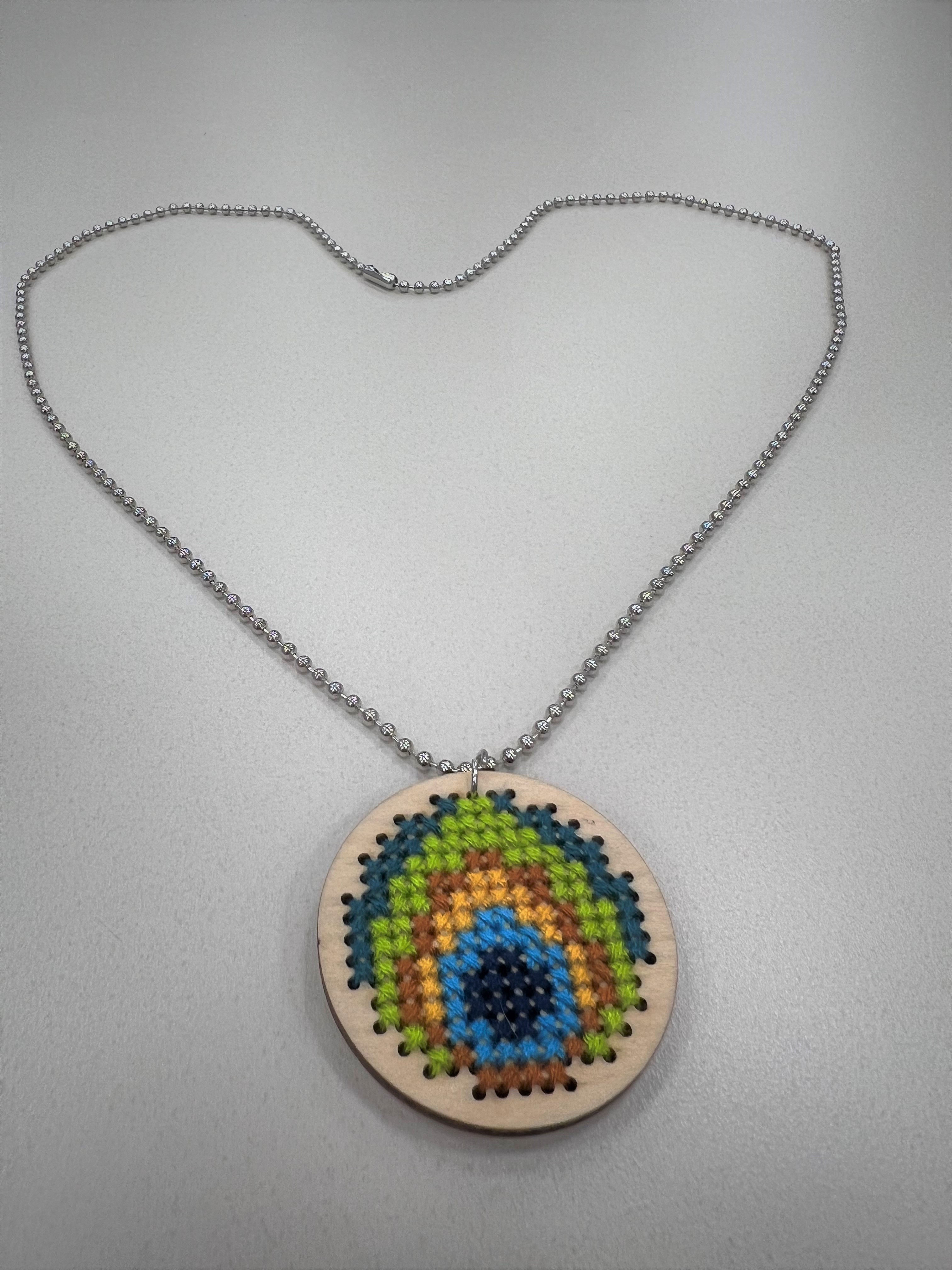 close up of a wooden pendant covered in a cross stitched peacock feather pattern