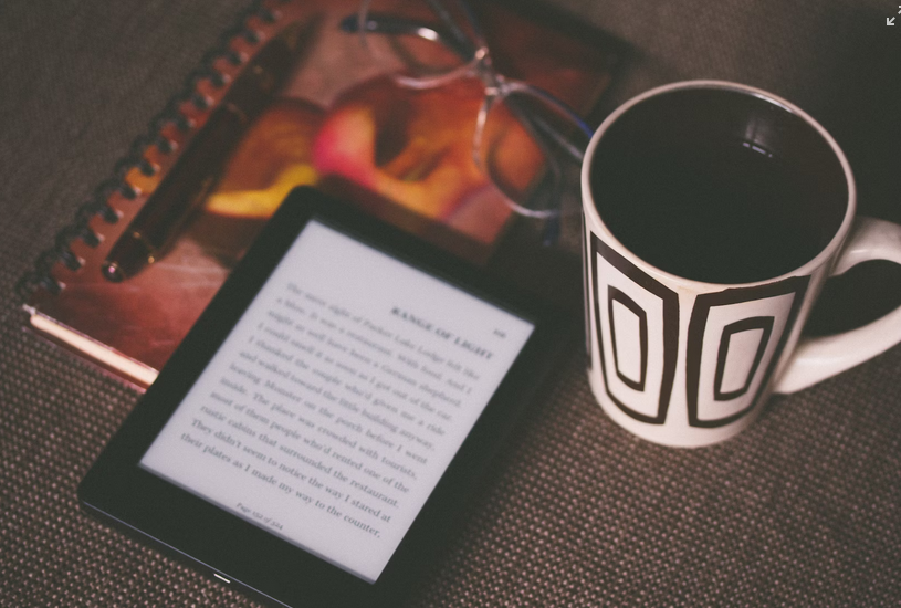 photo of an e-reader with out of focus text next to reading glasses, notebook, and mug of coffee