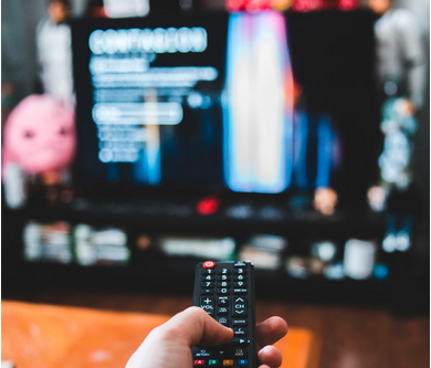 close up of a hand holding a tv remote; the background containing a TV is out of focus