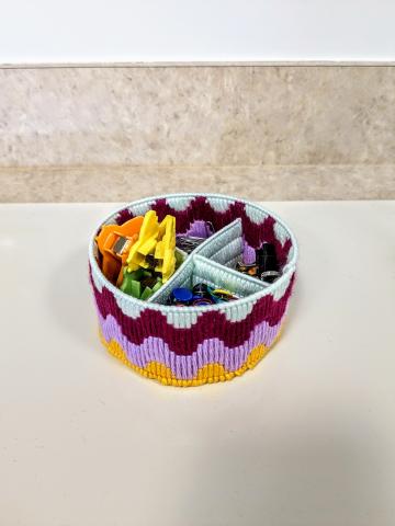 trinket tray made with pink, purple, yellow, and green yarn with push pins, binder clips, paper clips, and bag clips inside