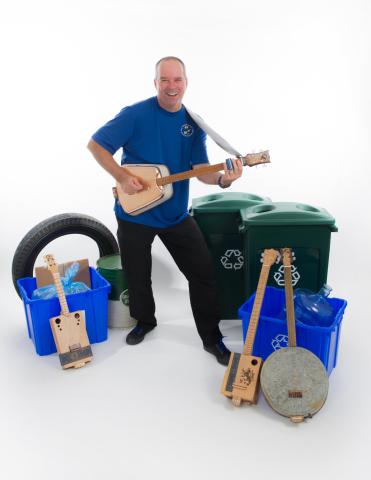 Tom, with Tom's Fun Band" is holding a guitar. Recycle bins, a tire, and other stringed instruments surround him on the ground.