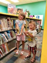 Two children standing by shelves of books, each holding a trophy.